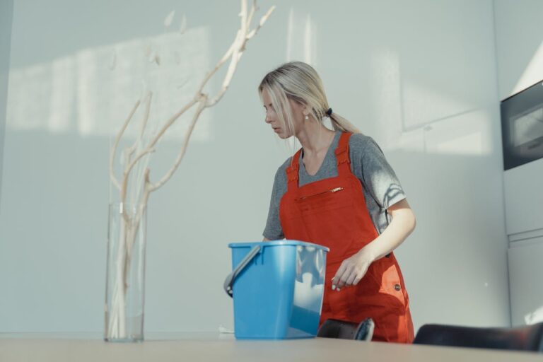 A woman in a red uniform cleans indoors with a blue bucket, ensuring a spotless environment.