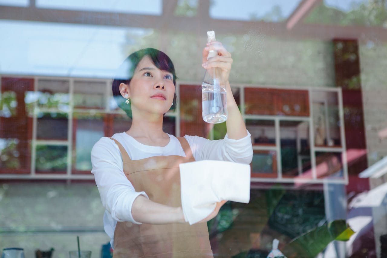 Asian woman wiping glass with a spray bottle and cloth, inside a restaurant setting.